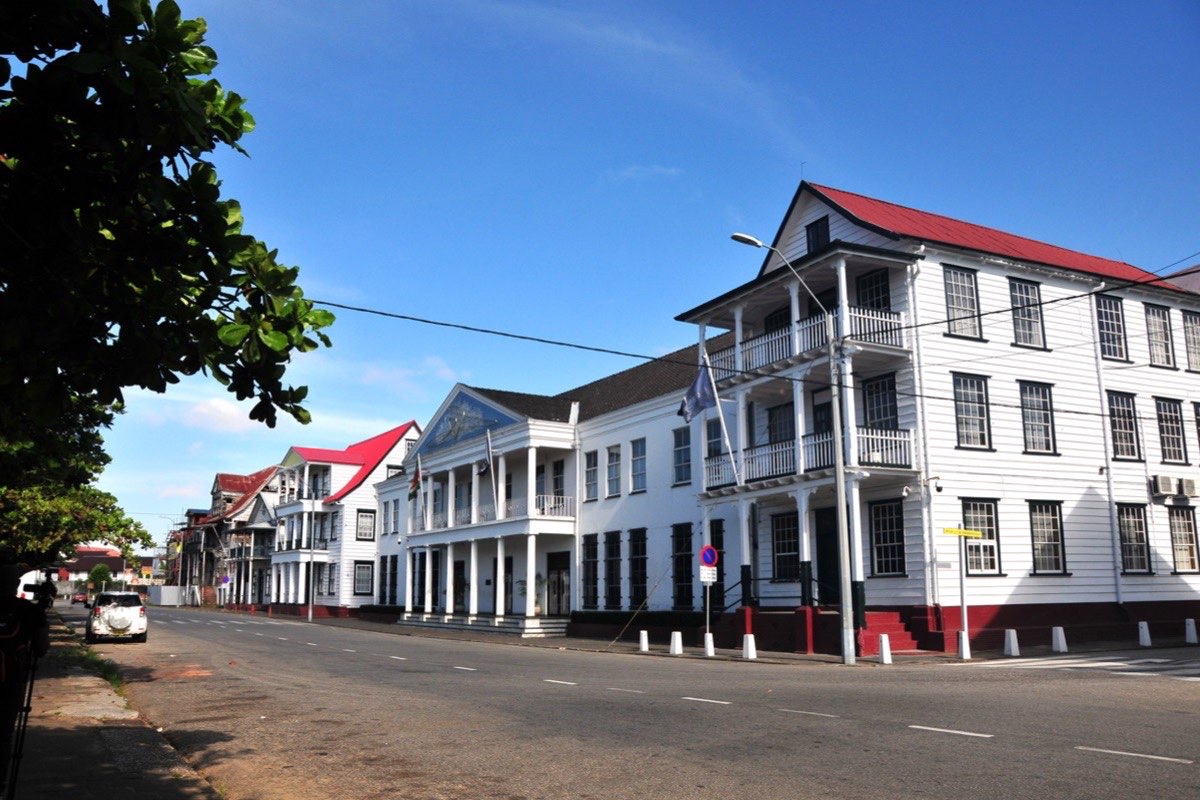 White timber colonial buildings with red roofs and colonnaded verandas lining the Waterkant, Paramaribo's riverfront street, under a clear blue sky.